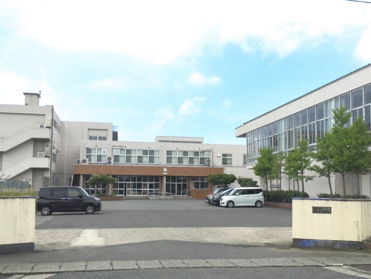 白い外壁の校舎とガラス窓のある泉小学校校舎の外観。門の奥に駐車場その奥に校舎が見えます。