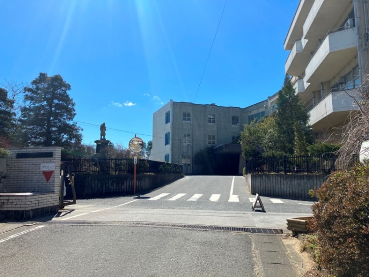 青い空の下、正門から見た学校の建物と横断歩道。学校の敷地内には銅像と植栽がある。