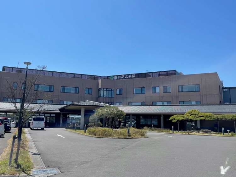 3階建ての池田記念病院の外観。周囲には駐車場と植栽が植えられています。