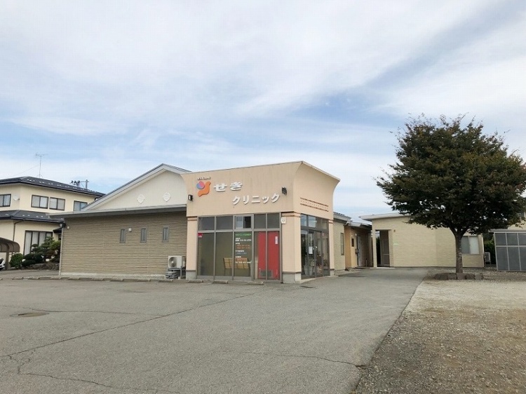 広い駐車場に面したせきクリニックの建物。左側には住宅が見えていて、前に大きな木が立っています。