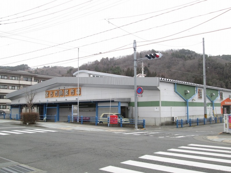青い柵と三角屋根が特徴的な宮古市魚菜市場の外観。背景には山が見えます。