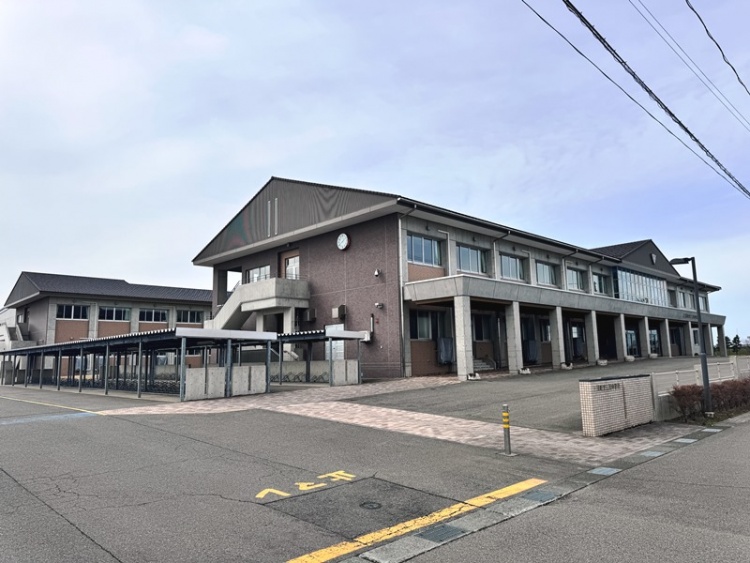 ブラウンの外壁を持つ2階建ての三川中学校校舎の外観。駐輪場があり、正面に広い舗装されたエリアがあります。