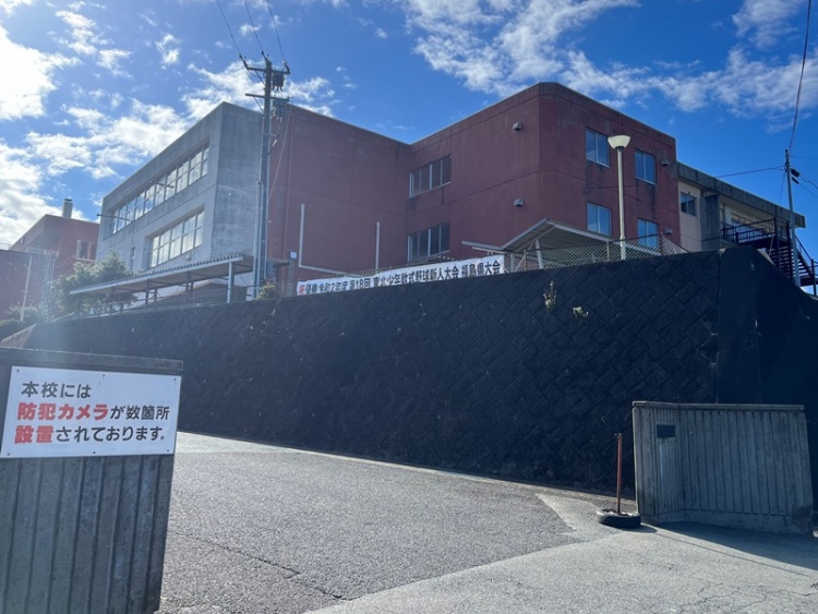 白い外壁と赤い屋根が特徴的な桜丘小学校の外観。手前には道路があり、門と校舎が見えます。