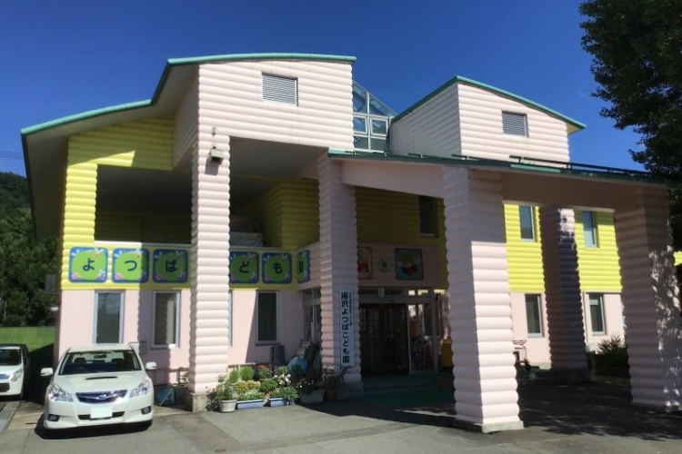 ピンクと黄色の外壁が特徴的なよつばこども園の外観。数台の車が停まっています。