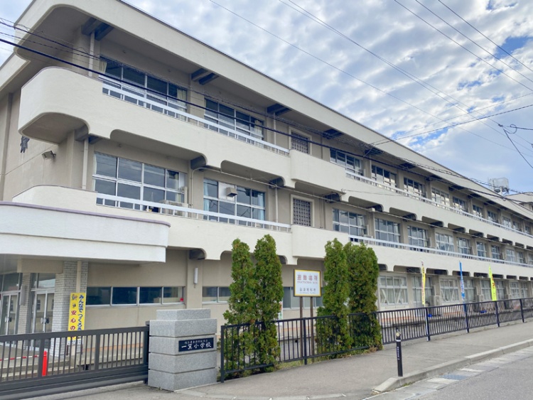 一箕小学校の校舎外観。3階建ての白っぽい建物で、正門近くに緑の植え込みがある。