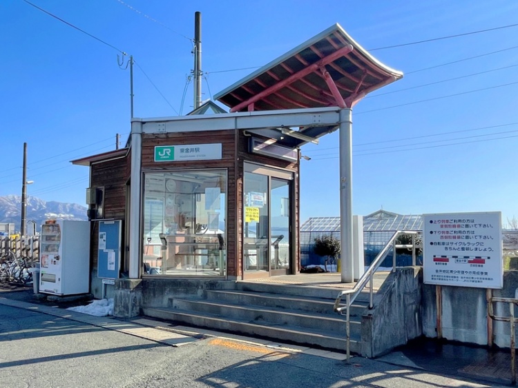JR東金井駅の小さな駅舎。自動販売機や掲示板が見え、背景には山々が広がっています。