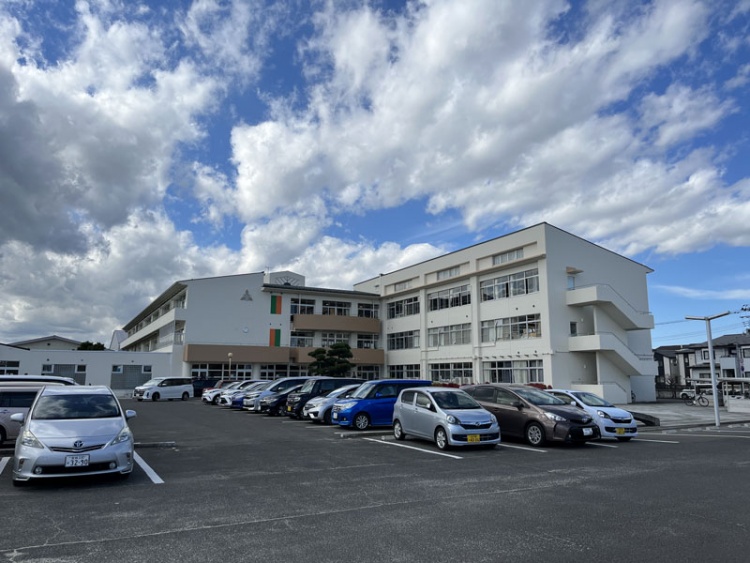白い外壁の名取市立第1中学校校舎の外観。駐車場には数台の車が停められています。