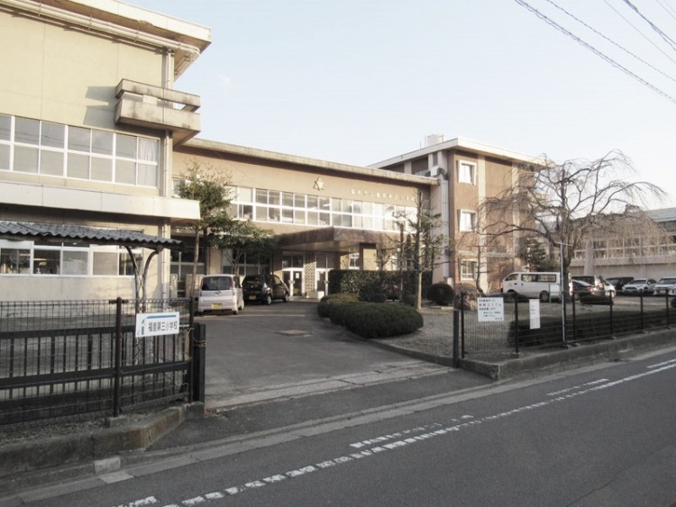 薄黄色の外壁が特徴的な福島第三小学校の校舎の外観。駐車場には車が停まっています。