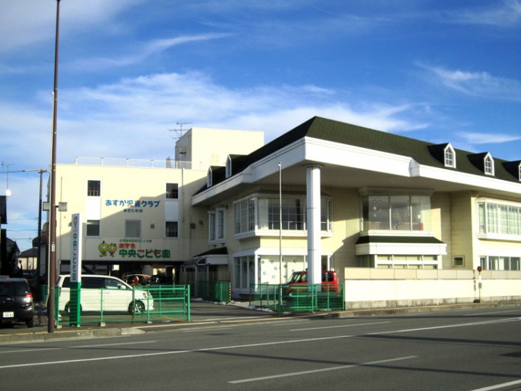 道路沿いにある大きな建物で、あきた中央こども園と書かれた看板が見えていて、建物の前に駐車場があり、車が停まっています。