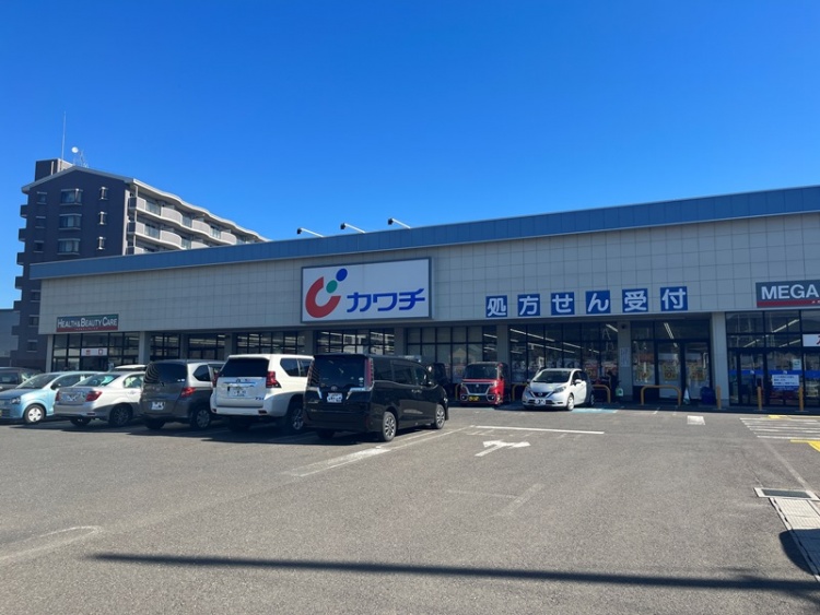 青の屋根が特徴的なカワチ薬品福島東店の外観。駐車場にはたくさんの車が停まっています。背景には建物が見えます。