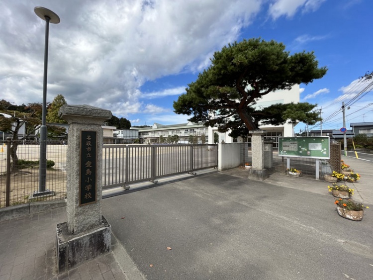 入口に石碑があり、松の木が立つ、名取市立愛島小学校校舎の外観。背景に校舎が見えます。