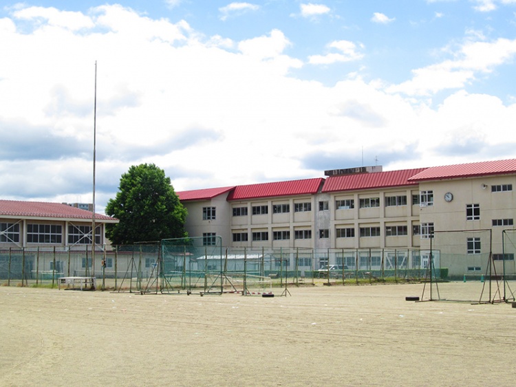 白い壁と赤い屋根が特徴的な下小路中学校校舎の外観。前面には広い運動場とフェンスが見えます。