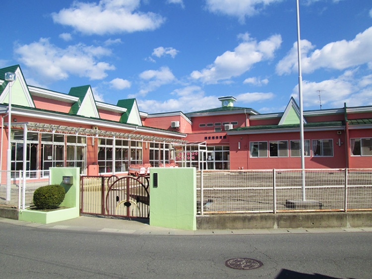 ピンク色の外壁に緑の屋根を持つ幼稚園の建物。門と柵が設置され、門の横に植木があります。