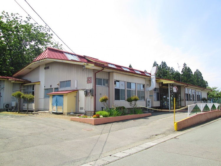 赤い屋根が特徴的なさくらがおか保育園の外観。複数の窓と出入口があり、周囲に緑の植栽が施されています。