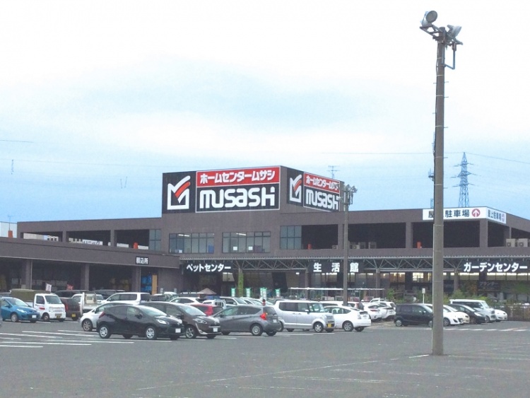 黒と白の外壁が特徴的なホームセンタームサシ酒田店の外観。駐車場には多数の車が駐車されている。ペットセンターや生活館、ガーデンセンターの看板が見えます。