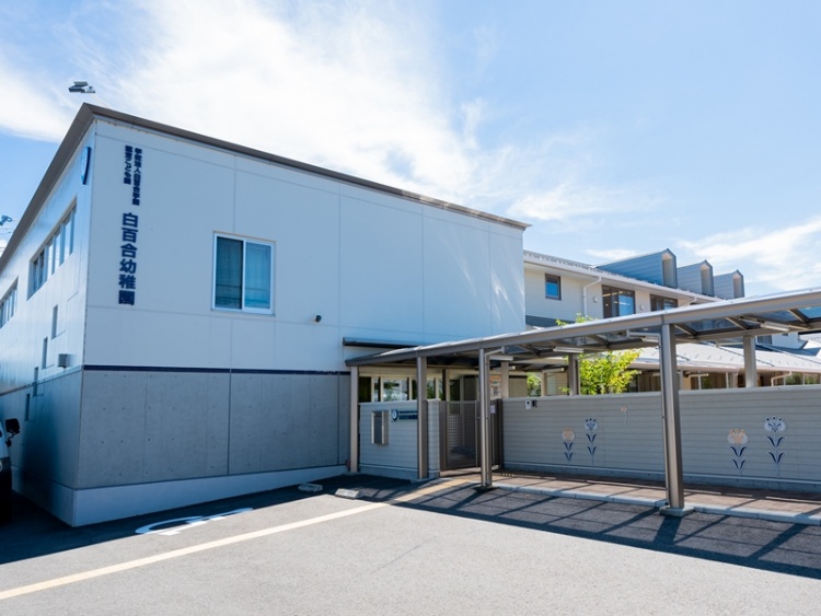 白とグレーの外壁が特徴的な二階建ての白百合幼稚園の外観。その前に設置された門やフェンスが見えます。