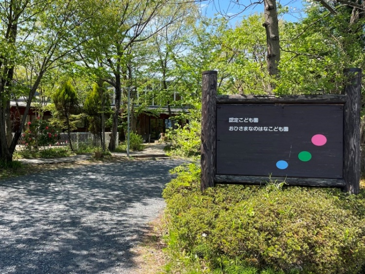 木立の中にあるおひさまなのはなこども園の外観。案内板には園の名前とカラフルなドットが描かれています。