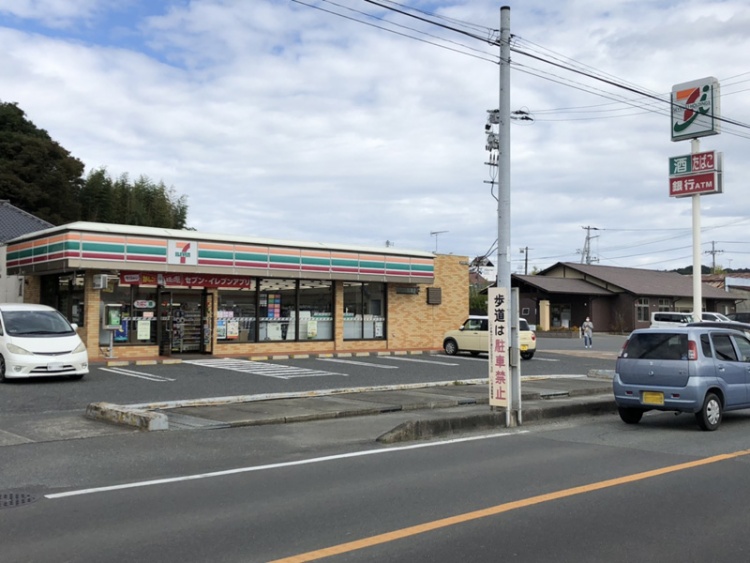 セブンイレブン岩城林城店の外観。駐車場が見えていて、手前に道路、標識柱には歩道は駐車禁止の看板があります。