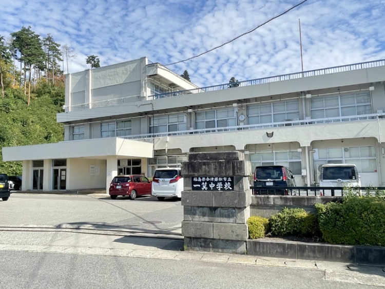 白い外壁が特徴的な一箕中学校の外観。門があり、駐車場には数台の車が停まっています。