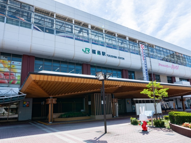 福島駅の西口の外観。駅名看板や建物の正面が写っていて、歩道には木が植えられ、キャラクター像があります。
