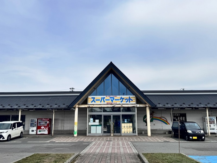 三角屋根が特徴的なスーパーマーケットラコスの外観。駐車場には数台の車が停められています。