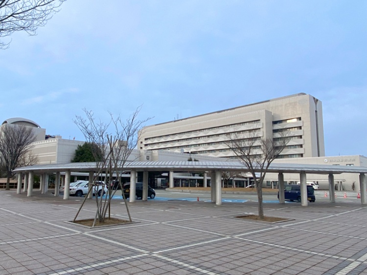 灰色の外壁の大きな日本海総合病院の外観。手前には駐車場と屋根付きの歩道があります。
