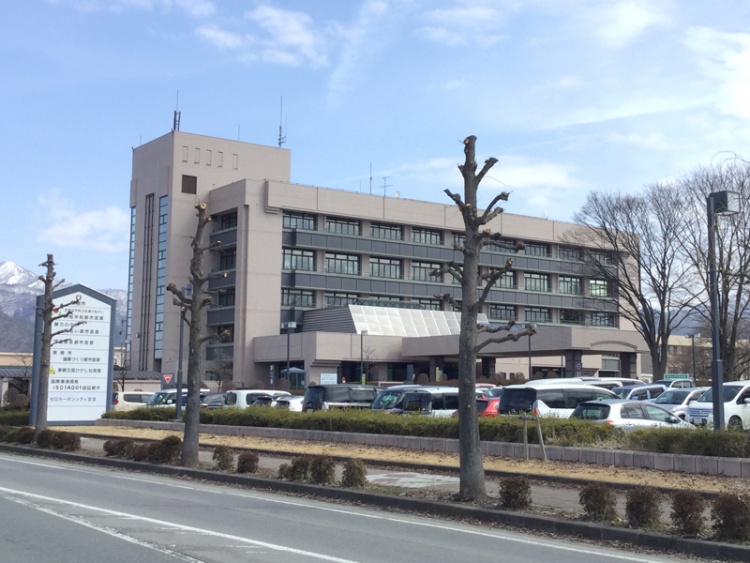 東根市役所の外観。駐車場には数台の車が停められています。案内看板が見え、遠くに山も見えます。