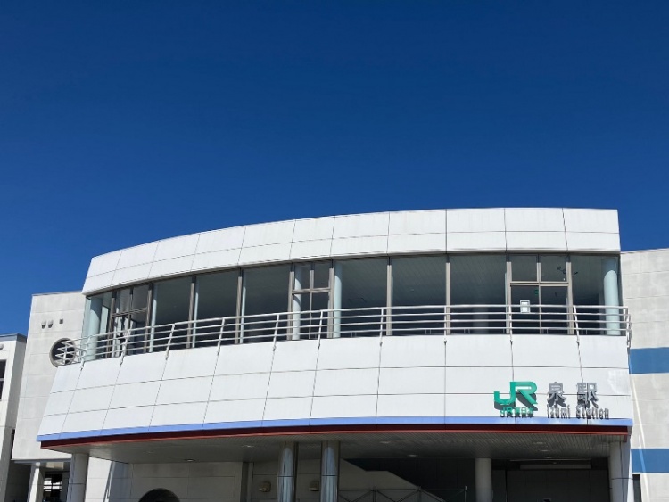 白い建物の壁に「JR泉駅」の文字が見える。曲面ガラスが特徴的なデザイン。青空が広がる。