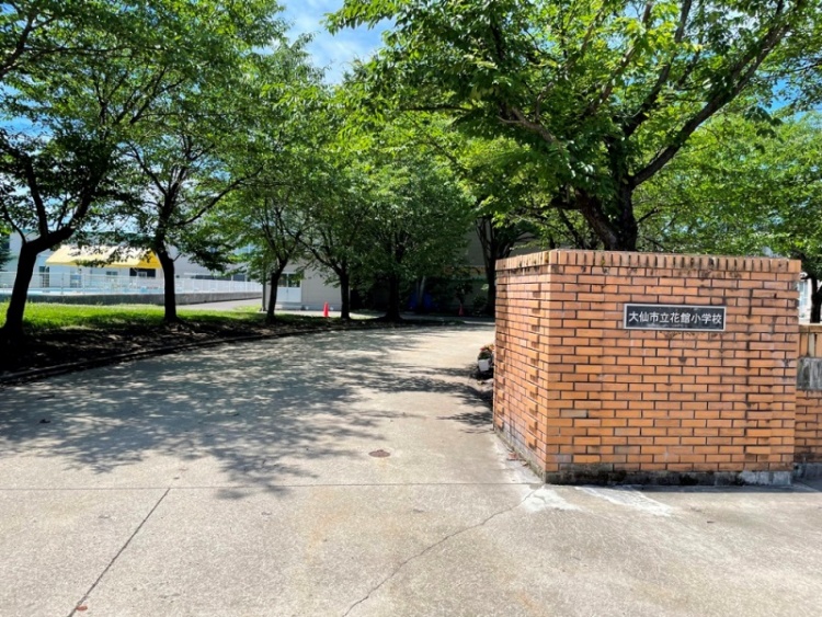 校門の前にレンガ造りの柱があり、大仙市立花館小学校の看板があり、周囲は木々に囲まれています。