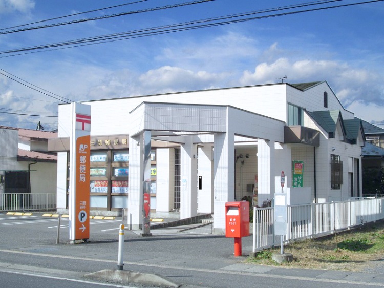 白い外壁とオレンジ色の看板がある三関郵便局の外観。手前に赤い郵便ポストと駐車場標識が見えます。