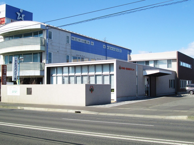 白と青の建物の前に茶色と白の平屋建てがあり、はちもり内科・消化器内科クリニックと書かれているクリニックの外観です。