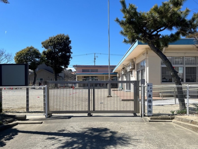 泉保育所の正門と校舎の一部。青空の下で、門の先に広場と建物が見えます。