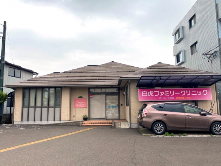 玄関とピンクの看板が特徴的な白虎ファミリークリニックの外観。駐車場には1台の車が停められています。