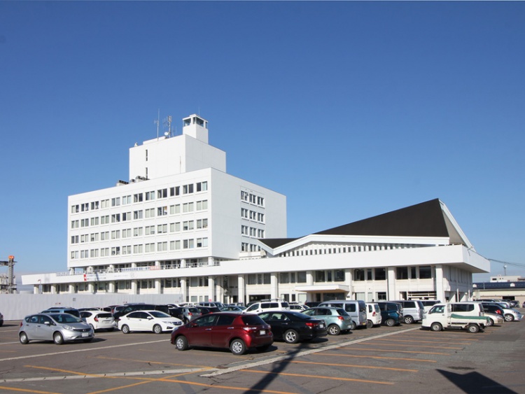 白い外壁の米沢市役所。手前は駐車場があり、数台の車が停められています。