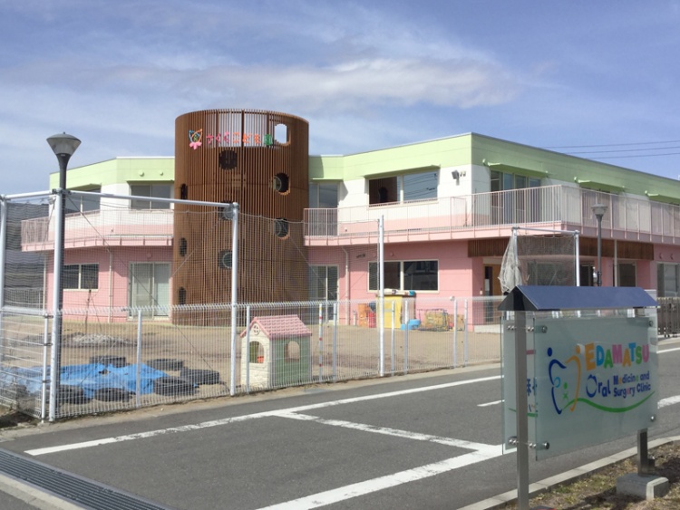 ピンクと緑の建物のさくらこども園の外観。フェンスで囲まれた遊び場と看板が見えます。