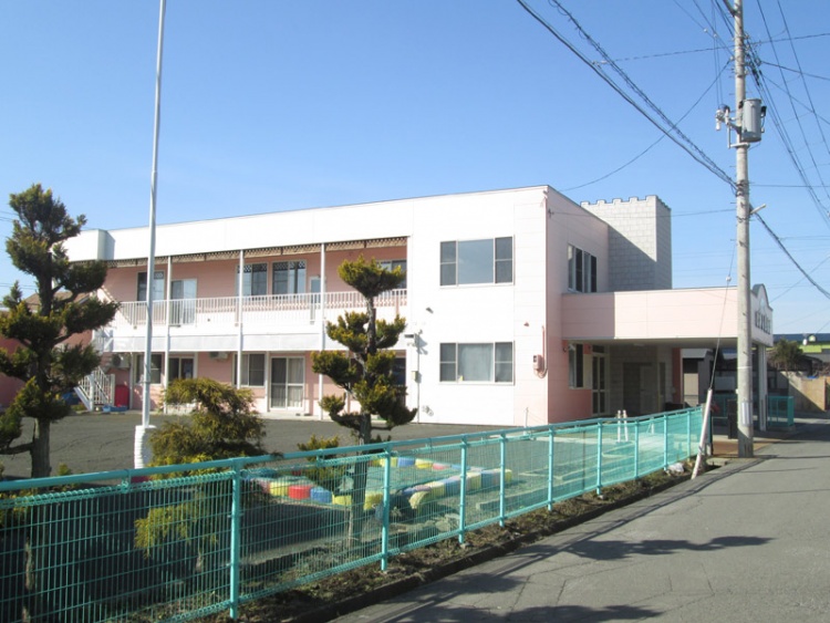 ピンク色の二階建てのひがし保育園の外観。前面にフェンスと遊具があります。