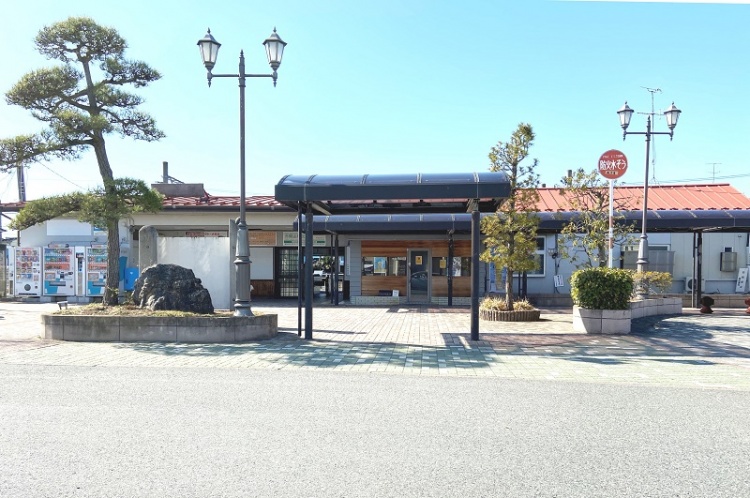 JR仙石線矢本駅の外観。緑の街路樹と石碑、屋根付きの通路が見えます。