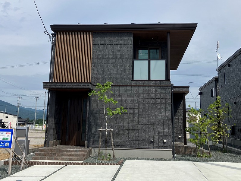 ダークグレーの外壁を持つ2階建ての住宅が、夕暮れの下に建っています。前には小さな庭があり、若木が植えられています。