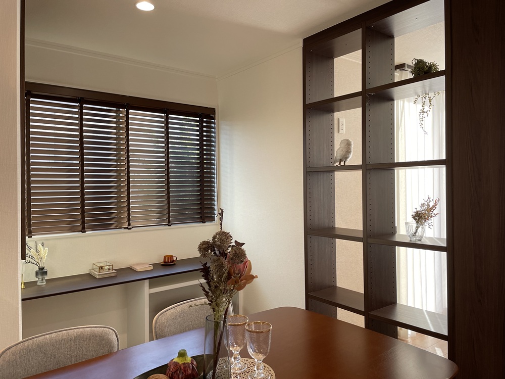ダークウッドの棚が空間を区切るリビングとダイニングです。窓からの光がブラインド越しに差し込み、棚には小物が飾られています。手前にはデスクと椅子があり、落ち着いた空間が広がっています。