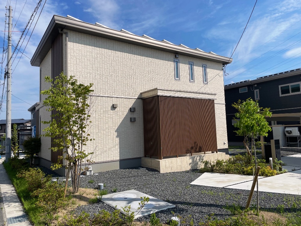 ベージュを基調とした外壁の2階建て住宅が、青空の下に建っています。植栽や小さな庭が整えられています。