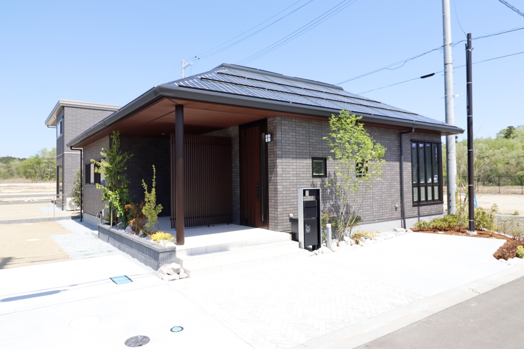 グレーの外壁と黒い屋根を持つ平屋建ての家が、日当たりの良い場所に建っています。玄関には木製の縦格子がアクセントとなり、周りには小さな植栽が植えられています。