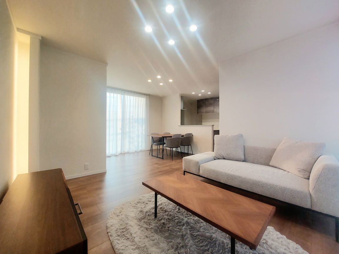 落ち着いた色合いのリビングダイニング。木目のフローリングに、ライトベージュのソファ、コーヒーテーブル、壁際にはローボードと照明付きの装飾コーナーがあり、間接照明で壁が柔らかく照らされている。