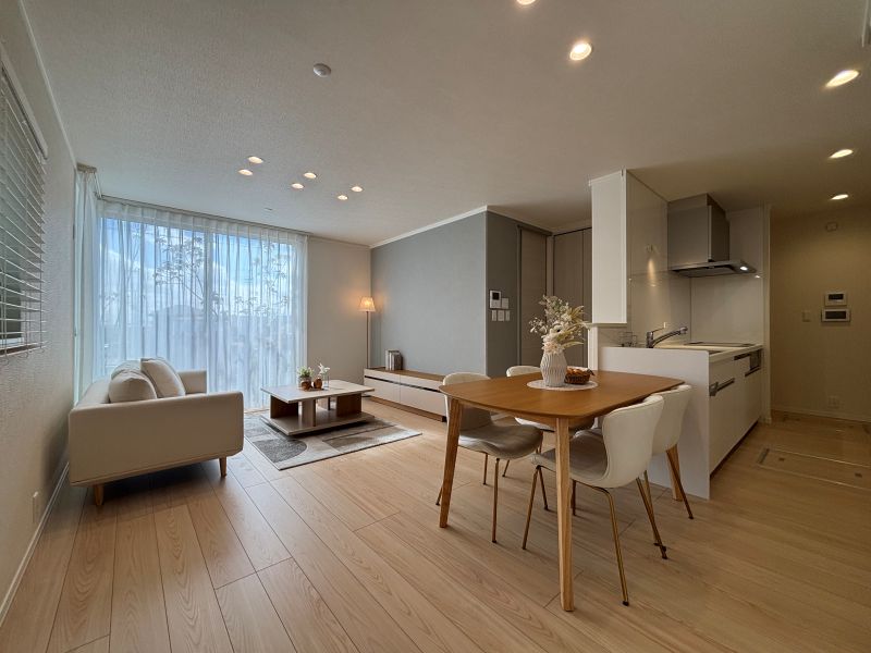 グレーのアクセントクロスと白を基調としたリビングダイニングキッチン。ソファ、ダイニングテーブル、椅子が配置されています。右側にはキッチンが見えます。