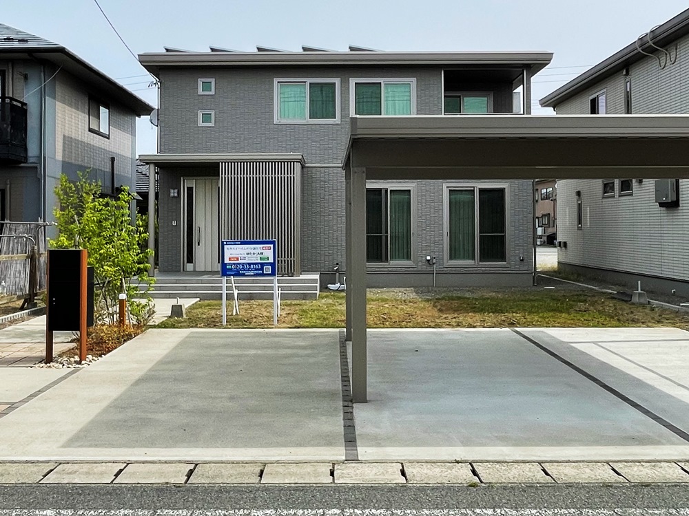 グレーを基調とした2階建て住宅の外観。手前にはカーポートがあり、2台分の駐車スペースがあります。庭には小さな木が植えられ、玄関前には看板が立っています。
