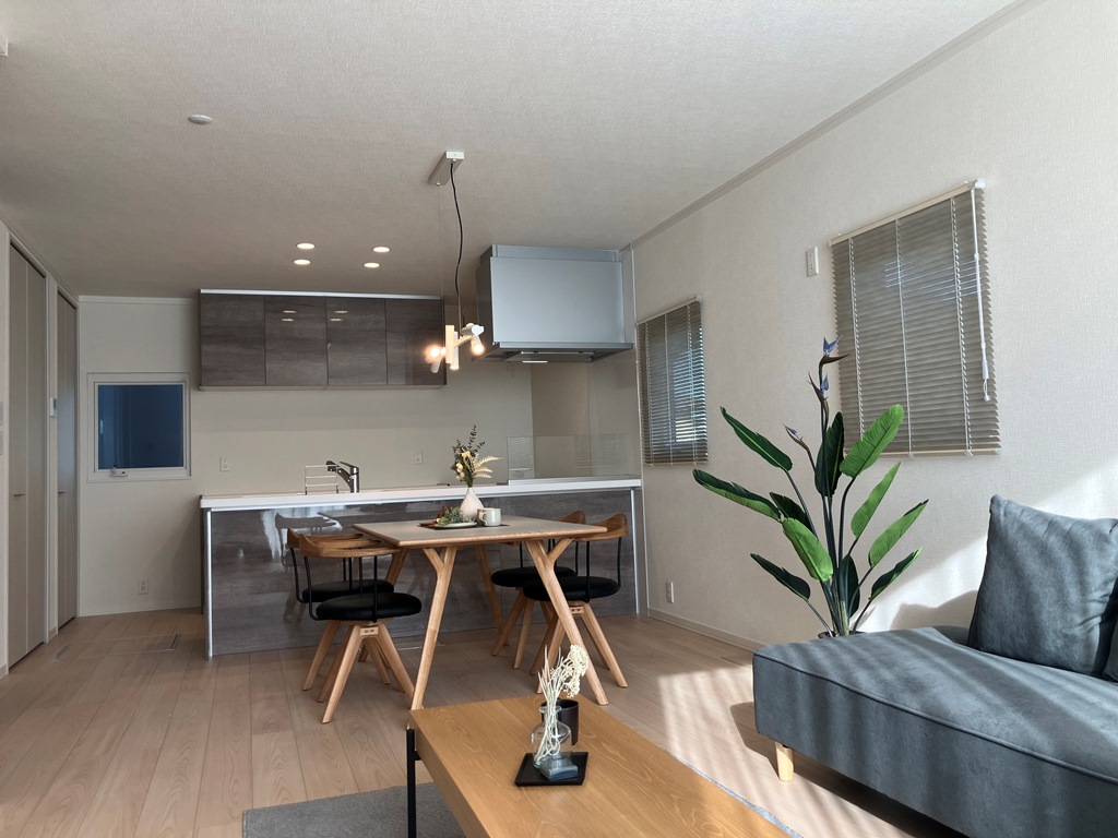 明るいリビングダイニングの風景。木製のダイニングテーブルと椅子が中央にあり、奥には対面式のキッチンが見える。右側には観葉植物とグレーのソファが置かれ、窓からの日差しが温かみを添えている。