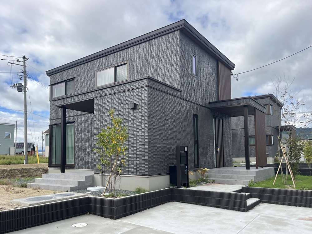 濃い外壁と縦格子のデザインが特徴的な二階建ての家で、整えられた植栽と段差のあるアプローチが重厚で落ち着いた印象を与えています。