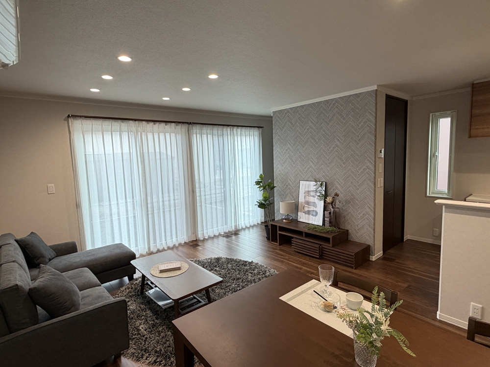 広々としたリビングダイニングに家具が配置されています。グレーのソファと丸いラグ、木製のコーヒーテーブルがあり、奥にはダイニングテーブルとキッチンが見えます。壁にはヘリンボーン柄のアクセントウォールがあります。窓からはたっぷりの光が差し込み、明るく落ち着いた雰囲気の空間です。