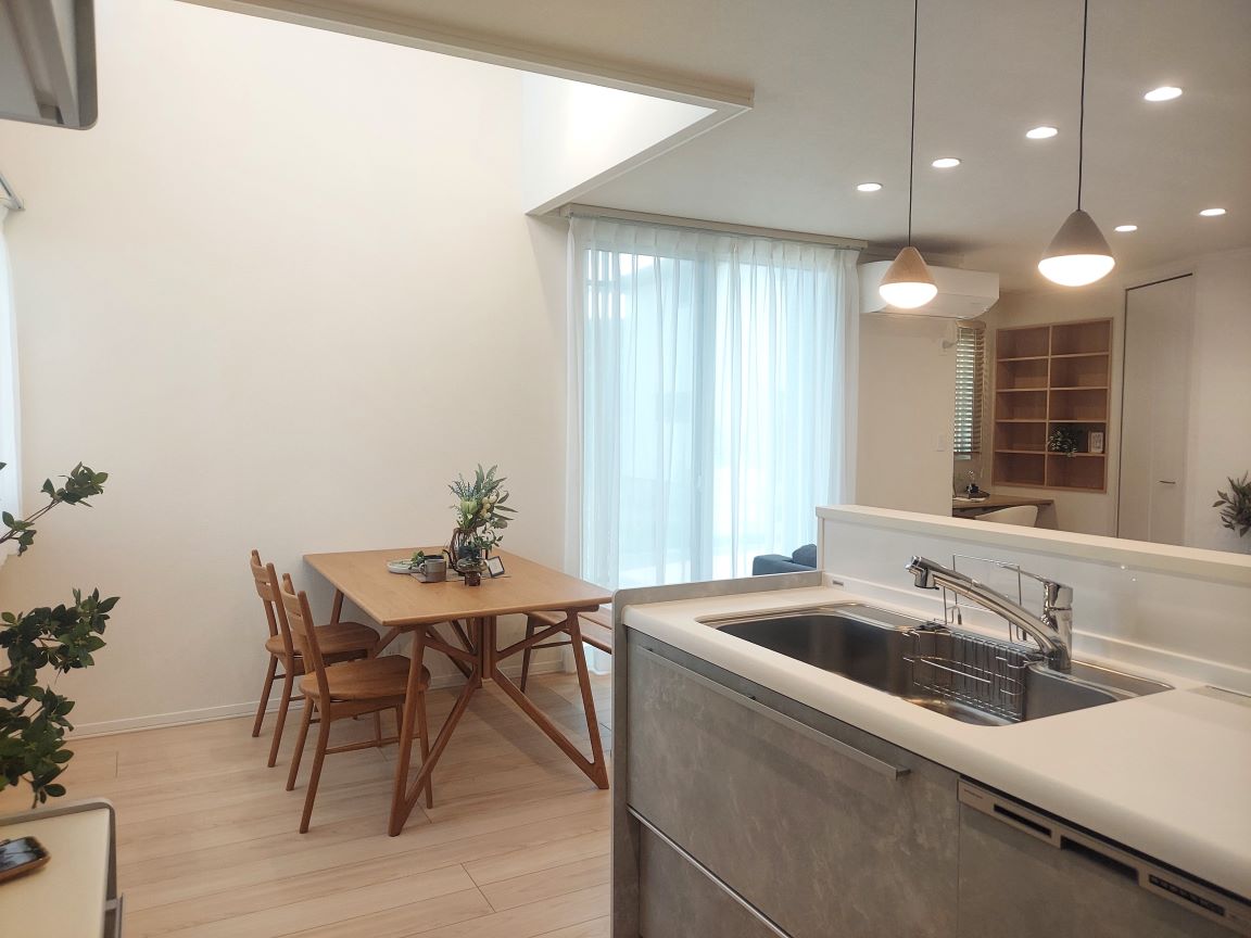 ナチュラルな木目調のフローリングの上に木製のダイニングテーブルと椅子が置かれたダイニングエリア。奥にはブルーグレーのソファを置いたリビングがあり、大きな窓から光が差し込む明るい空間。天井にはダウンライトが設置されている。