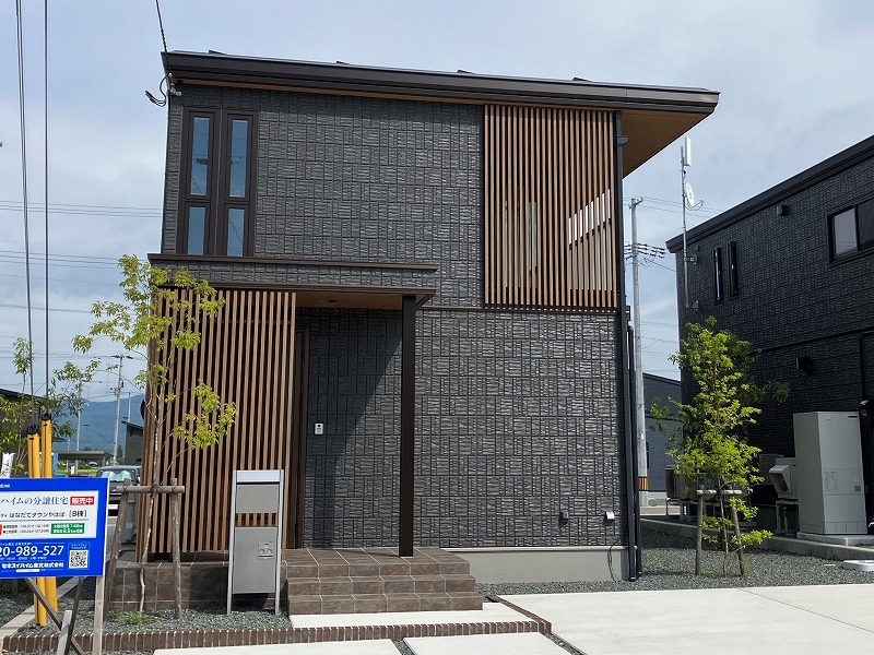 ダークグレーの外壁と木目調の縦格子が特徴的な2階建ての住宅が、夕暮れの下に立っています。玄関前には階段があり、小さな木々が植えられています。’