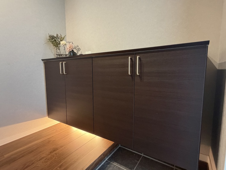 木目調の玄関カウンター収納には、植物やアートフレームが飾られています。キャビネットの下にある間接照明が、柔らかい光を放ち、落ち着いた雰囲気を演出しています。